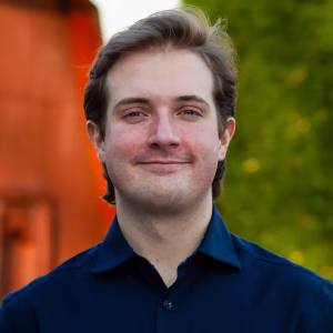 A young man with light brown hair and a slight smile stands outdoors, wearing a dark blue shirt. The background features greenery and a blurred structure